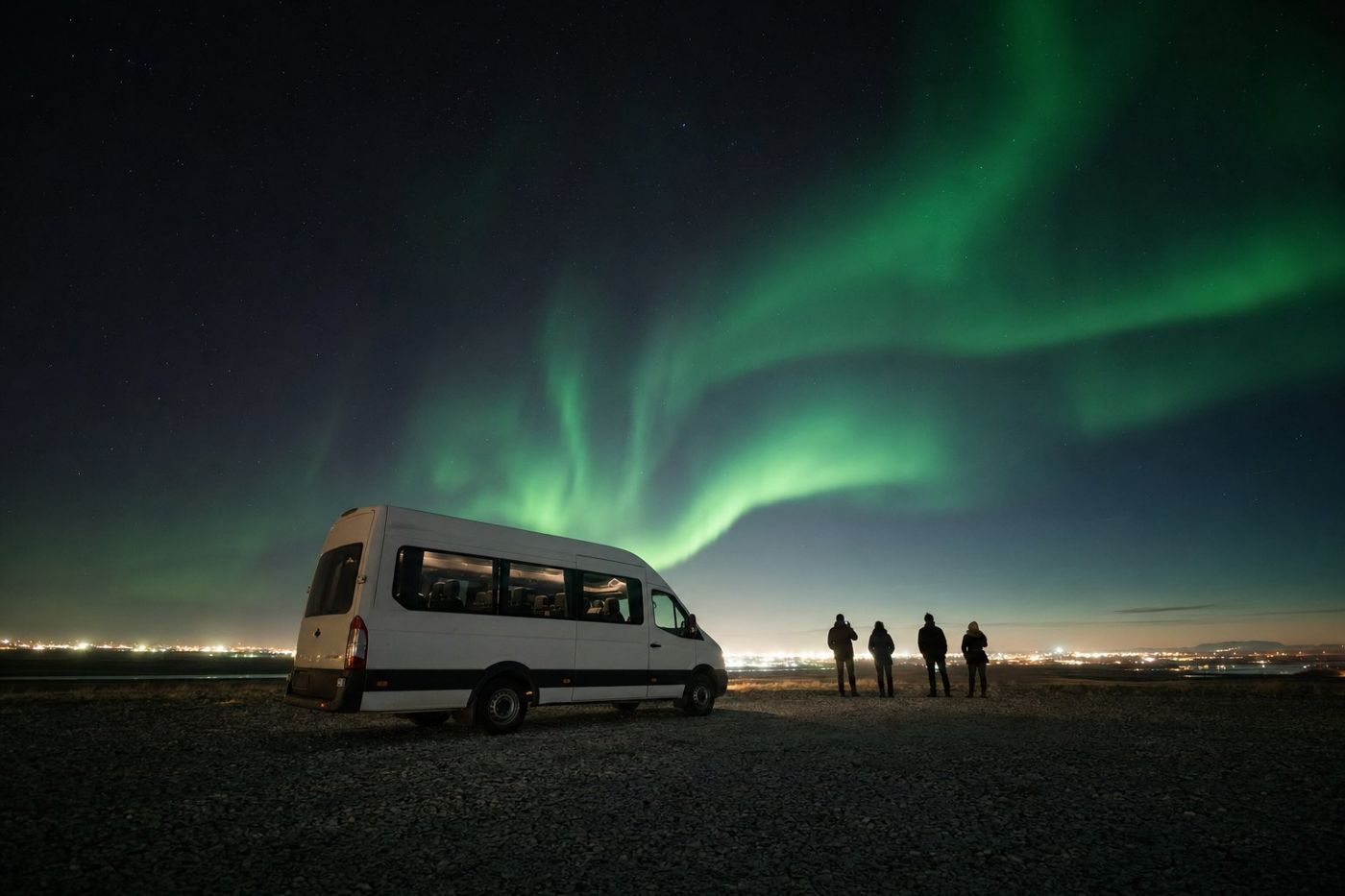 Un minibus arrêté sur une route de gravier sombre avec une aurore verte au-dessus et la lueur de Reykjavik à l'horizon derrière