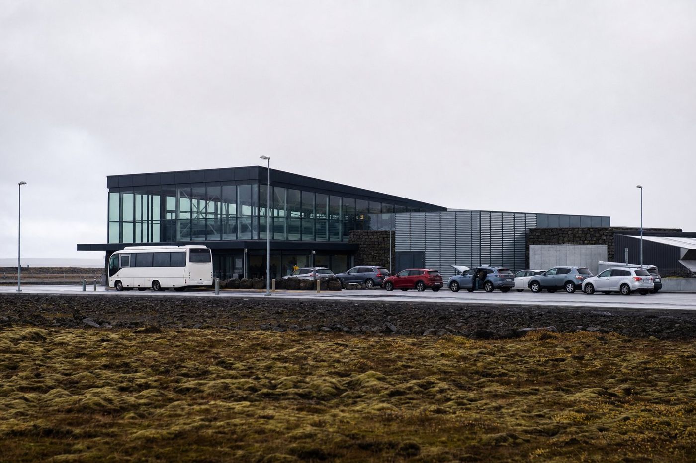 Un terminal d'aéroport moderne sur une plaine de lave plate, avec une navette et des voitures garées devant